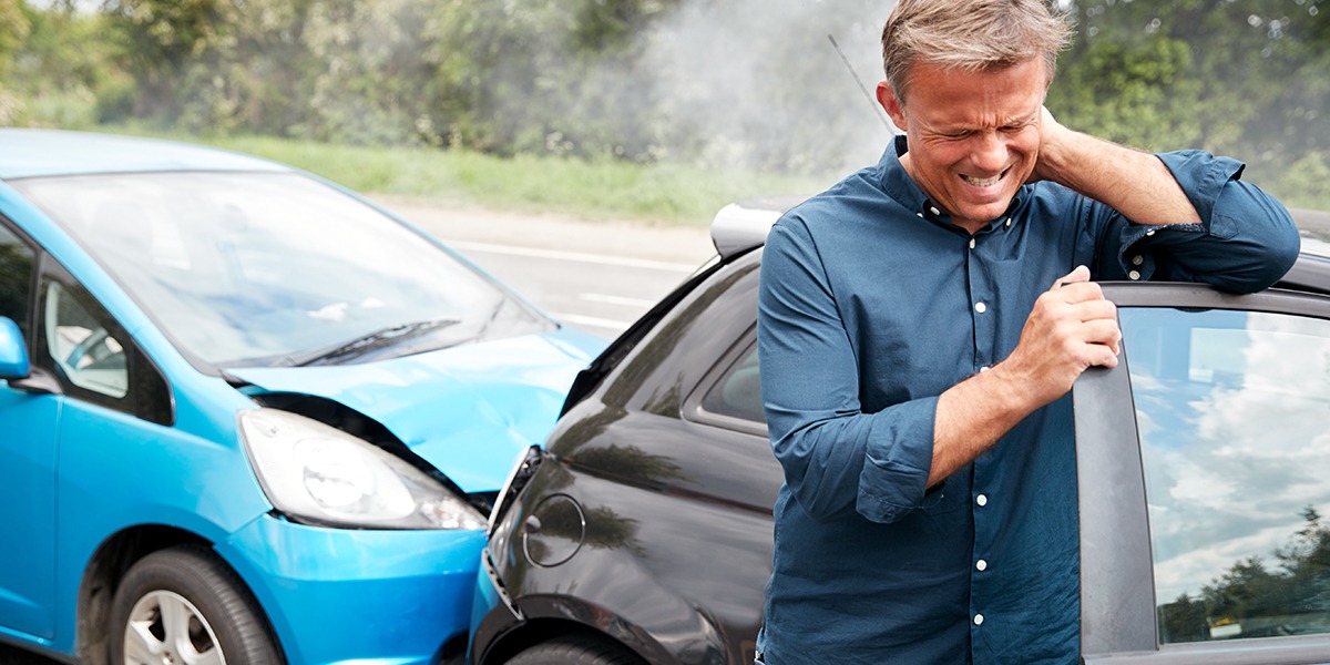 Man in a blue shirt grimaces while leaning on a dented black car after a collision with a blue car in the background.
